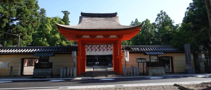 豊満神社四脚門と緑の木々の写真。朱色と緑のコントラストが美しい